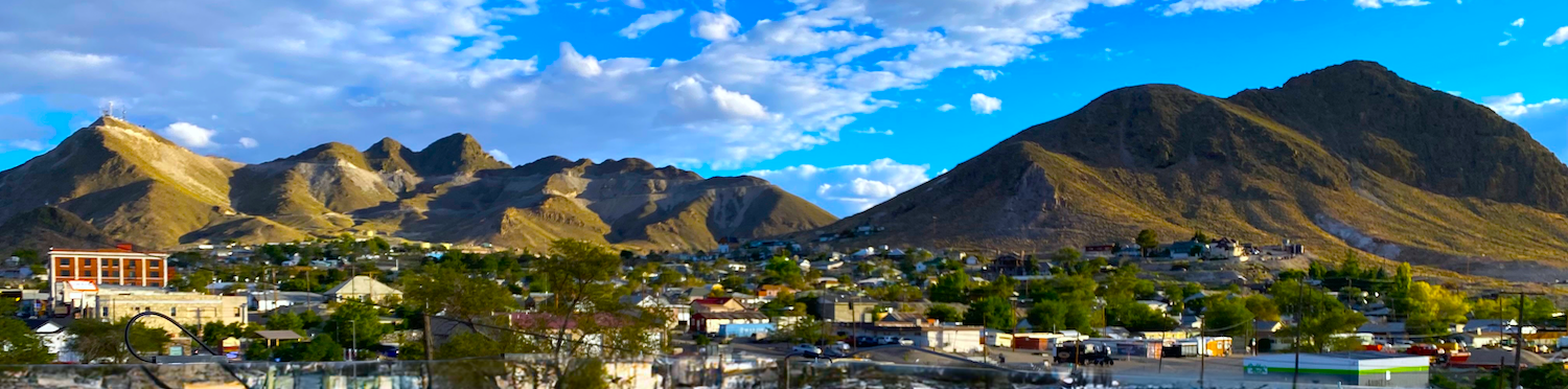 Desert town with deciduous trees, downtown area, neighborhoods, mountains in background, blue sky with fluffy clouds. This is the city skyline of Tonopah, NV.